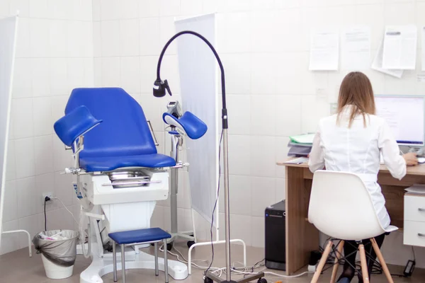 Baksida syn på blond kvinna gynekolog doktor i vit Uniform i klinik sjukhus fungerar på datorn. Gynecological skåp med stol på bakgrunden. Kvinna hälsa, nyfödda och graviditet Concept — Stockfoto