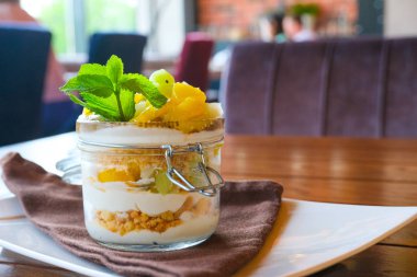 Cam kavanozda sağlıklı katmanlı meyve tatlısı. Kivi, mango, ananas, şeftali, chia tohumu, mascarpone, krem şanti, nane ve granola ahşap masada. Arka planda bulanık restoran. Tatlı menü konsepti