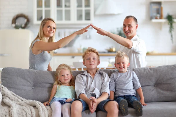 Kanepede oturan üç çocuk ile mutlu tam aile, anne ve baba başları üzerinde elleri ile çatı figürü yapma. Yeni bina konut ev satın daire ve konut kavramı.