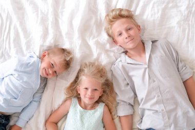 Üç neşeli sarışın çocuk yatakta birlikte yatıyor, kameraya bakıyor ve gülümsüyorlar. İki erkek ve kız. Mutlu aile, erkek ve kız kardeş