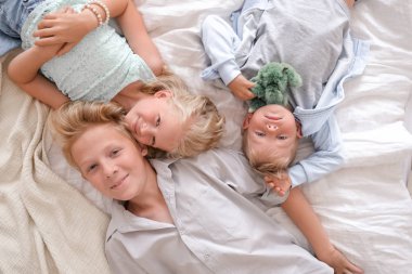Üç neşeli sarışın çocuk yatakta birlikte yatıyor, kameraya bakıyor ve gülümsüyorlar. İki erkek ve kız. Mutlu aile, erkek ve kız kardeş