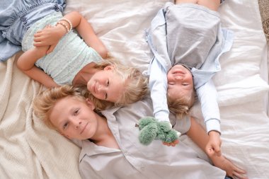 Üç neşeli sarışın çocuk yatakta birlikte yatıyor, kameraya bakıyor ve gülümsüyorlar. İki erkek ve kız. Mutlu aile, erkek ve kız kardeş