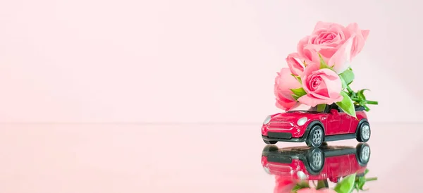 Red toy car delivering bouquet of pink rose flowers on pink background. Banner. Place for text. Mirror reflection. February 14 card, Valentine\'s day. Flower delivery. 8 March, Happy Women\'s Day.