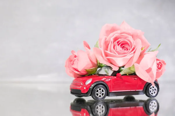 Red toy car delivering bouquet of pink rose flowers on grey background. Mirror reflection. Place for text. February 14 card, Valentine day. Flower delivery. 8 March, International Happy Women Day.