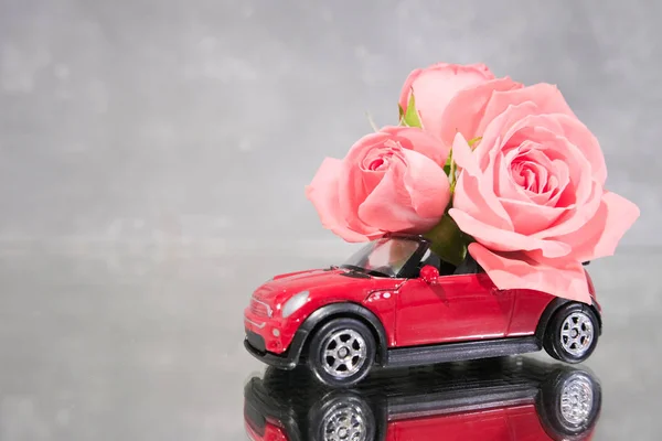 Red toy car delivering bouquet of pink rose flowers on grey background. Mirror reflection. Place for text. February 14 card, Valentine day. Flower delivery. 8 March, International Happy Women Day.