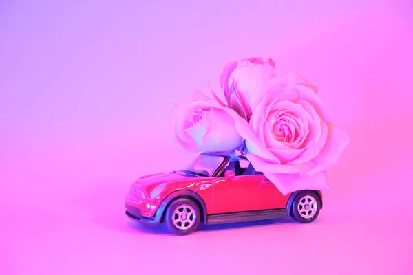 Red toy car delivering bouquet of pink rose flowers on trendy color neon background. Place for text. February 14 card, Valentine\'s day. Birhday. 8 March, International Happy Women\'s Day