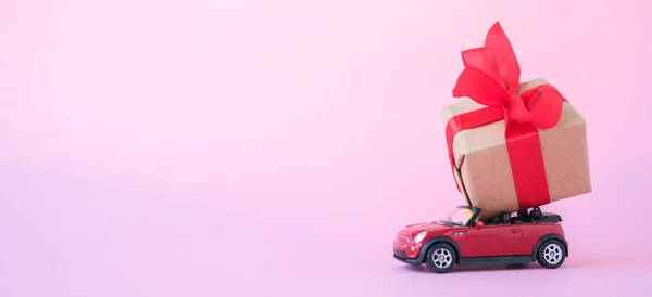 Банер з червоним іграшковим автомобілем, що постачає подарункову коробку з червоною стрічкою на рожевому тлі. Місце для тексту. Продажі, День народження, різдвяний час — стокове фото