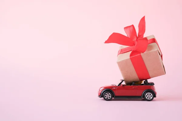 red toy car delivering gift box with red ribbon on pink background. Place for text. Sales, Birthday, christmas time. St.Petersburg Russia 16 jul 2019.