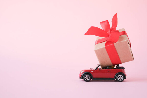 red toy car delivering gift box with red ribbon on pink background. Place for text. Sales, Birthday, christmas time. St.Petersburg Russia 16 jul 2019.