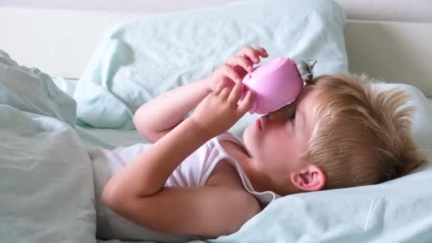 En liten blond pojke ligger i sängen och ler, spelar väckarklocka tidigt på morgonen. Pojken lyssnar när klockan tickar. Dags att vakna — Stockvideo