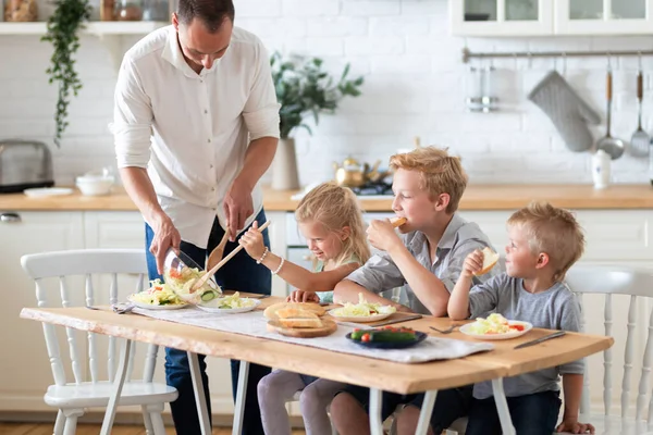 family father with three kids two sons and daughter eating healthy food in kitchen at home, dad puts green salad on plates to his kids