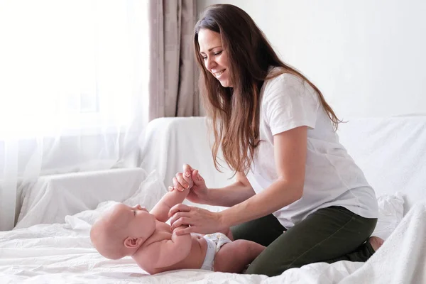 Jovem mulher branca mãe faz massagem para bebê infantil feliz na cama branca em casa. Babycare, esporte e maternidade feliz . — Fotografia de Stock