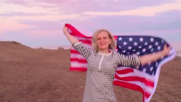 4k. gelukkig glimlachende vrouw in de zomer kleding met nationale VS vlag buiten oceaan zonsondergang - Amerikaanse vlag, land, patriottisme, onafhankelijkheidsdag en mensen concept. — Stockvideo