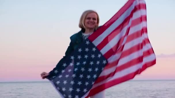 4k. χαρούμενη χαμογελαστή γυναίκα με καλοκαιρινά ρούχα με εθνική σημαία των ΗΠΑ σε εξωτερικούς χώρους ηλιοβασίλεμα του ωκεανού - Αμερικανική σημαία, χώρα, πατριωτισμός, ημέρα ανεξαρτησίας και οι άνθρωποι έννοια. — Αρχείο Βίντεο