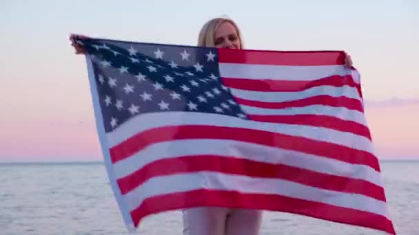 4k. gelukkig glimlachende vrouw in de zomer kleding met nationale VS vlag buiten oceaan zonsondergang - Amerikaanse vlag, land, patriottisme, onafhankelijkheidsdag en mensen concept. — Stockvideo