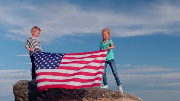 4K. Menino e menina loira acenando bandeira nacional dos EUA ao ar livre sobre o céu azul no verão bandeira americana, país, patriotismo, dia da independência 4 de julho . — Vídeo de Stock