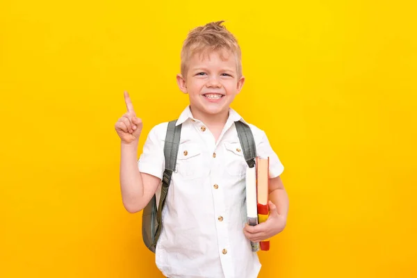 Terug naar school. Portret van kleine blonde jongen basisschool met tas en boeken in wit shirt. Wijzende vinger omhoog. Gele studio achtergrond. Onderwijs. Kijken en glimlachen naar de camera. — Stockfoto