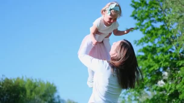 4kだ。母は娘を青空に高く持ち上げる。緑の芝生の上で一緒に夏に公園で楽しい屋外遊びをしている幸せなお母さんと女の子。家庭生活. — ストック動画