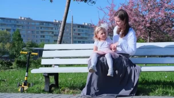 4kだ。母と娘は市内公園の白いベンチに座って夏に話しています。家族の生活様式。一緒に時間,母性. — ストック動画