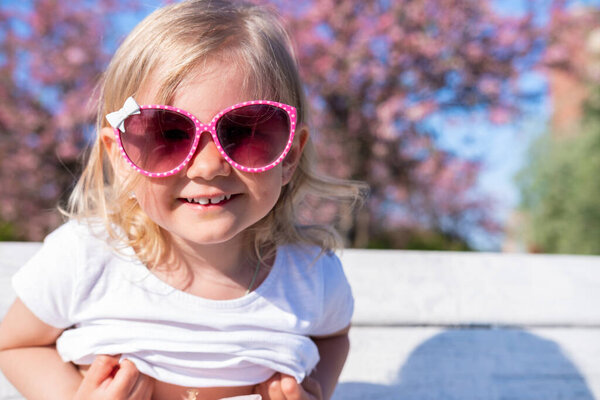 Cute little baby girl smiling in pink eyeglasses at summer, looking at camera. Happy blonde caucasian child playing having fun outdoor. 