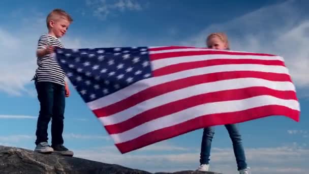 4K. Menino e menina loira acenando bandeira nacional dos EUA ao ar livre sobre o céu azul no verão bandeira americana, país, patriotismo, dia da independência 4 de julho . — Vídeo de Stock