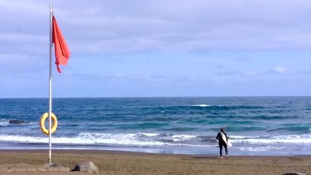 Vista trasera Hombre surfista con tabla de surf está caminando a lo largo del océano con grandes olas. Bandera roja de advertencia ondeando con fuerte viento . — Vídeos de Stock