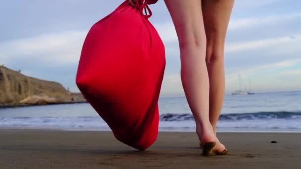 Vánoční dívka Santa Claus chůze na písečné pláži oceánu s velkou červenou taškou plnou dárků - Nový rok dovolenou a cestování. Zpomalený pohyb. — Stock video