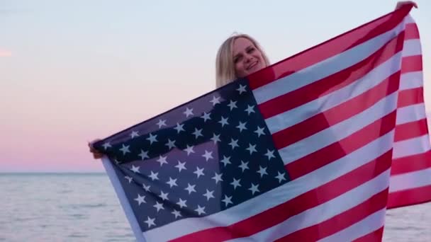 4k. gelukkig glimlachende vrouw in de zomer kleding met nationale VS vlag buiten oceaan zonsondergang - Amerikaanse vlag, land, patriottisme, onafhankelijkheidsdag en mensen concept. — Stockvideo