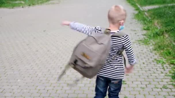 4k blonde boy playing on street, spinning with backpacks after school. Child wearing mask safety coronavirus. — Stock Video