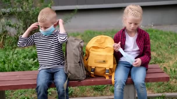 4k torna a scuola. Ragazza e ragazzo i bambini si siedono a parlare sulla panchina vicino alla scuola mantenendo la distanza sociale indossando maschera — Video Stock