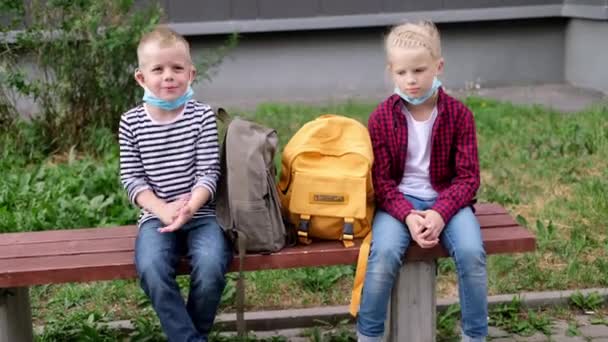 4k terug naar school. Meisje en jongen kinderen zitten te praten op bank in de buurt van de school houden sociale afstand dragen masker — Stockvideo