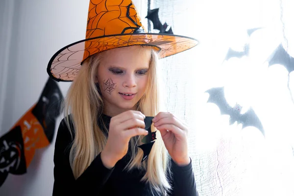 Cadılar Bayramı için hazırlanan cadı kostümlü mutlu kız siyah yarasaların odadaki pencerelerini süslüyor. — Stok fotoğraf