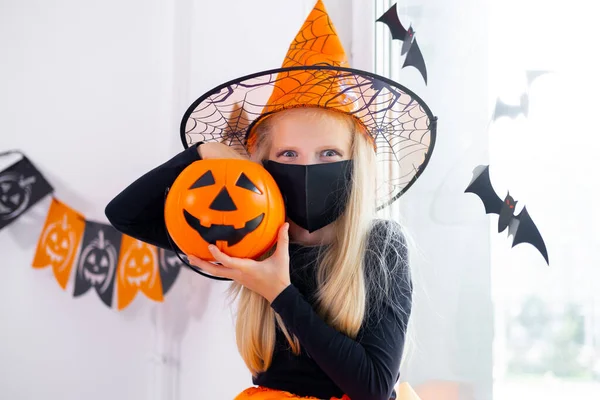 Retrato chica rubia en traje de bruja con máscaras faciales con cubo de caramelo en pandemia coronavirus preparándose para Halloween protegiendo de COVID-19 —  Fotos de Stock
