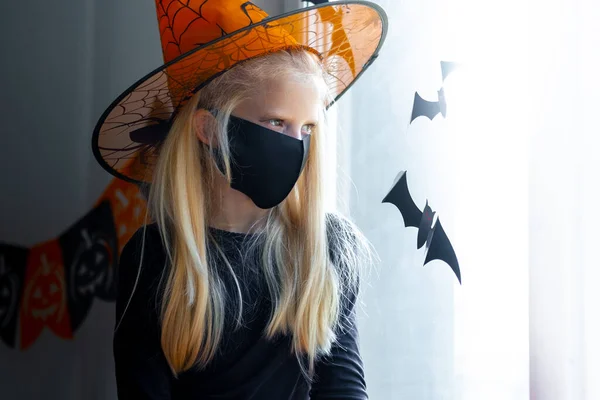 Chica rubia triste en traje de bruja con máscaras en la cara mirando en la ventana en casa. preparándose para Halloween protegiéndose del coronavirus —  Fotos de Stock