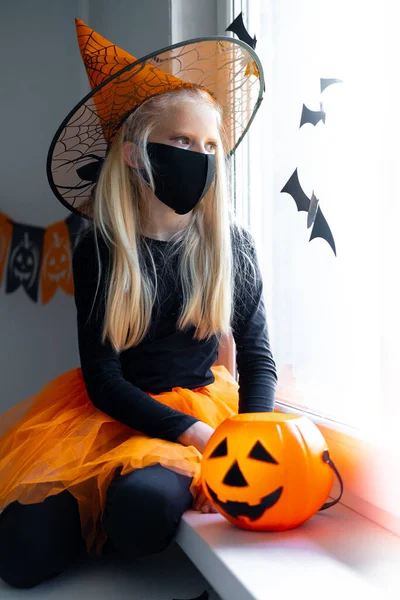 Sad thoughtful blonde girl in witch costume wearing mask looking in window at home with candy bucket. preparing for Halloween protecting from covid — Stock Photo, Image