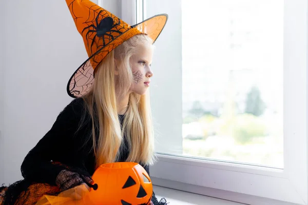 Sorglig omtänksam blond flicka i häxdräkt tittar in genom fönstret hemma med godis hink. Förberedelser för Halloween skyddar mot coronavirus. — Stockfoto