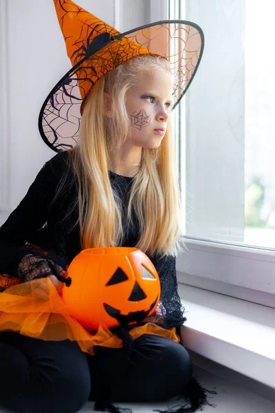 Θλιβερό ξανθό κορίτσι με στολή μάγισσας κοιτάζοντας στο παράθυρο στο σπίτι με τον κουβά καραμέλα. προετοιμασία για το Halloween προστασία από coronavirus. — Φωτογραφία Αρχείου