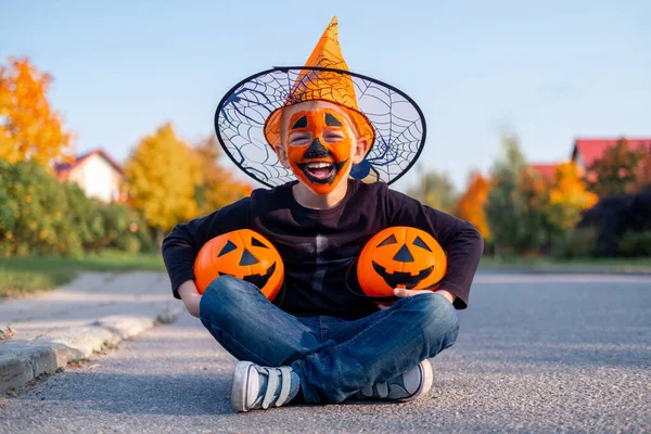 Niños de Halloween. Chico risueño con mascarilla de calabaza en sombrero de bruja con dos baldes de caramelo sentado en la calle —  Fotos de Stock
