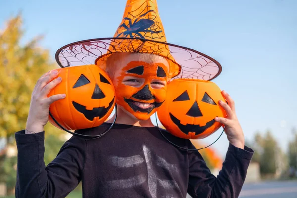 Niños de Halloween. Chico risueño con mascarilla de calabaza en sombrero de bruja con dos baldes de caramelo sentado en la calle —  Fotos de Stock