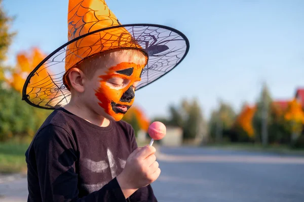 Niños de Halloween. niño con mascarilla de calabaza en traje de bruja sombrero comiendo dulces de cubos sentados en la calle —  Fotos de Stock