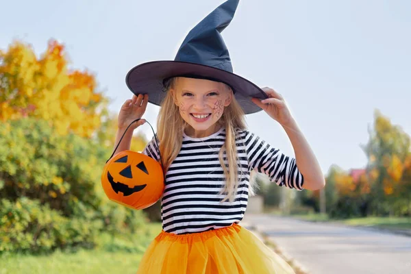 เด็กฮาโลวีน รูปสาวที่ยิ้มในหมวกแม่มด กับถังขนมฟักทอง เด็กตลกในชุดคาร์นิวัลกลางแจ้ง — ภาพถ่ายสต็อก