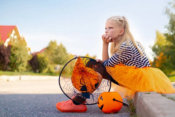 Niños de Halloween. Chica triste triste infeliz en sombrero de bruja con cubo de caramelo de calabaza sentado solo en la calle en trajes de carnaval al aire libre —  Fotos de Stock