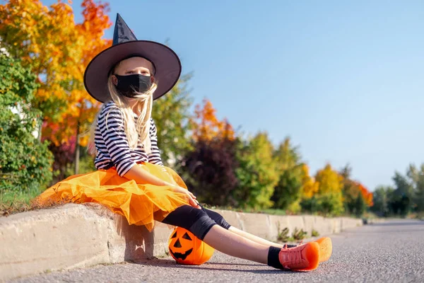 Halloween kids mask. Portrait blonde girl in witch costume with pumpkin bucket. Child wearing black face masks outdoors protecting from COVID-19