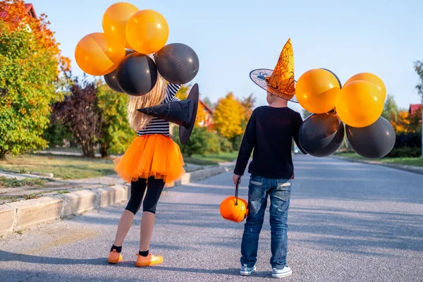 Miúdos do Halloween. Voltar vista irmão e irmã em trajes de carnaval ao ar livre. Rapaz e rapariga de chapéu de bruxa com baldes de doces de abóbora. Crianças engraçadas com balões laranja e preto. — Fotografia de Stock