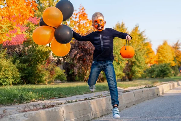 Niño de Halloween en trajes de carnaval celebrando Halloween al aire libre. Niño con mascarilla de calabaza y cubos de caramelo corriendo por caramelos con globos naranjas y negras. —  Fotos de Stock