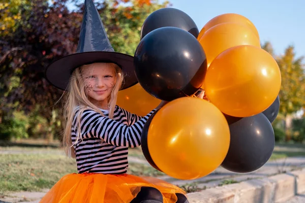Cadılar Bayramı çocukları. Cadı şapkalı, turuncu ve siyah balonlu gülümseyen kız. Karnaval kostümlü komik çocuklar. — Stok fotoğraf