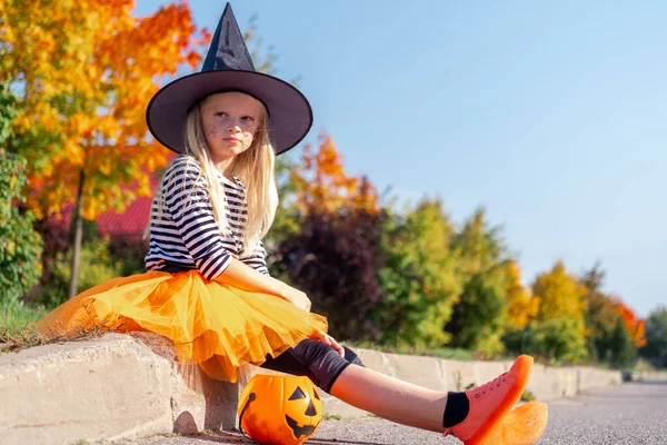 Niños de Halloween. Chica triste triste infeliz en sombrero de bruja con cubo de caramelo de calabaza sentado solo en la calle en trajes de carnaval al aire libre —  Fotos de Stock