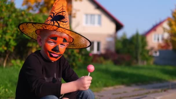 Halloween kinderen. jongen met pompoen masker in heksenkostuum hoed eet snoep van emmers die op straat zitten. Langzame beweging — Stockvideo