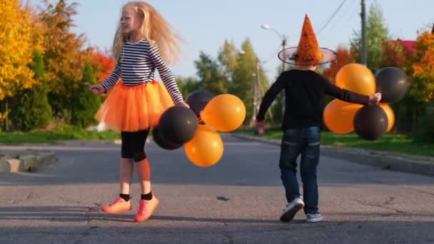 Langzame beweging. Halloweenkinderen. Broer en zus in carnaval kostuums buiten. Jongen en meisje hebben plezier met oranje en zwarte ballonnen. — Stockvideo