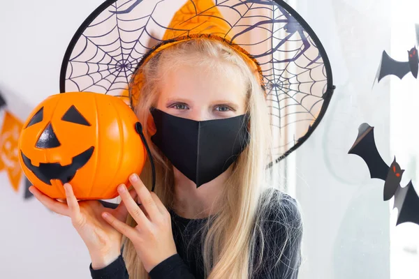 Retrato chica rubia en traje de bruja con máscaras faciales con cubo de caramelo en pandemia coronavirus preparándose para Halloween protegiendo de COVID-19 —  Fotos de Stock